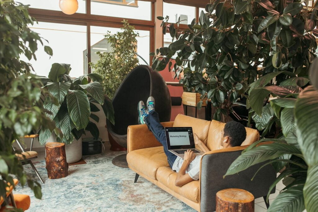Man relaxing on couch, working on laptop in plant-filled room.