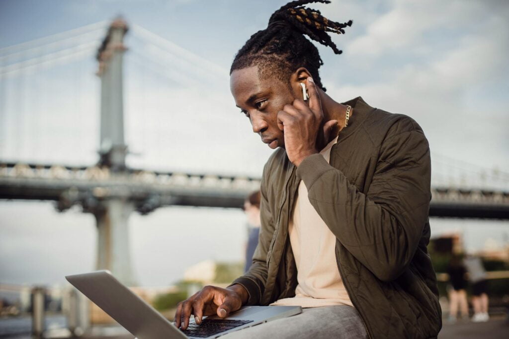 Man using laptop and earphones near bridge outdoors.